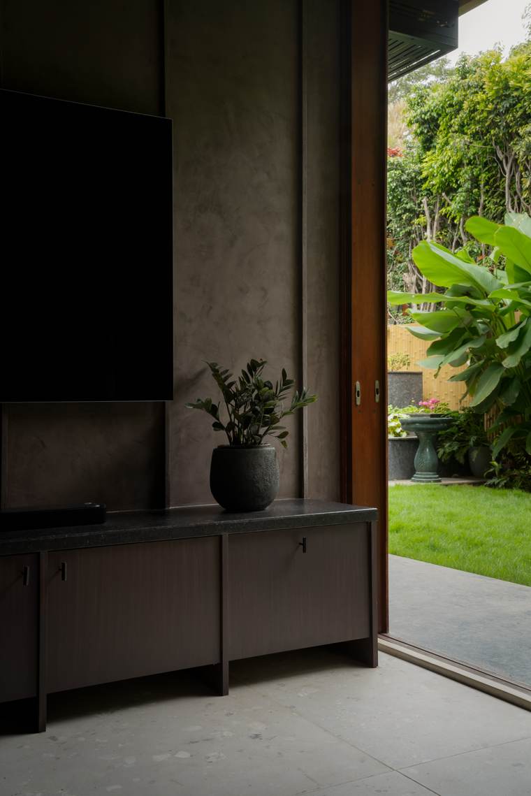 Dark plastered TV wall with a small textured pot holding a plant on a black stone-topped timber unit, beside an open sliding door to the garden with banana leaves and a birdbath