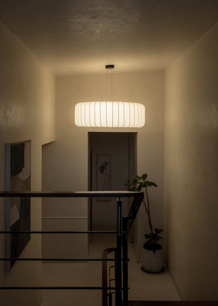 Staircase landing lit by a large pleated drum pendant, with a large abstract artwork, a passage beyond and a rubber plant behind a black railing