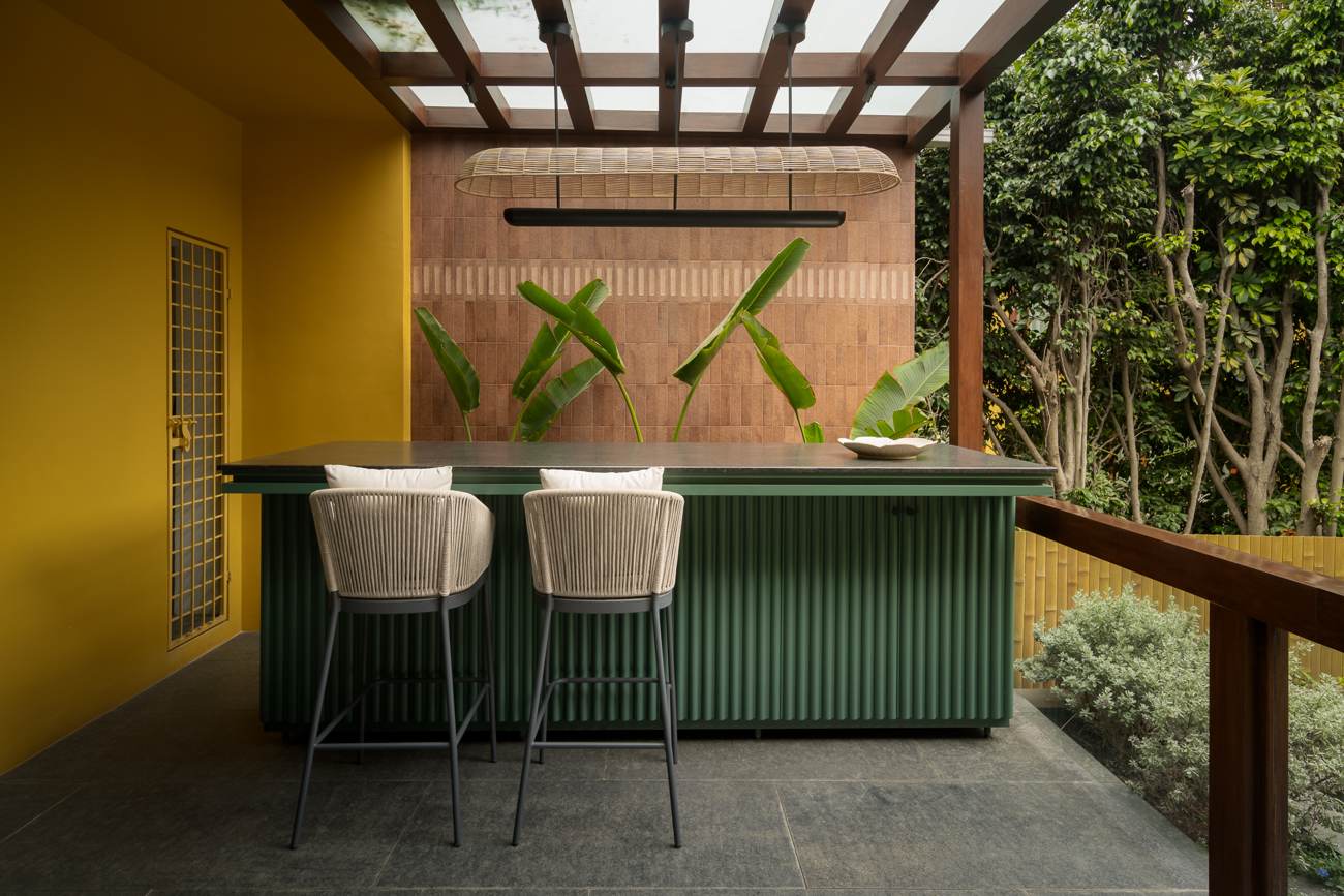 Terrace bar with a green fluted counter, two rope-backed stools, a woven pendant over a terracotta tile wall, banana leaves, a yellow wall and garden beyond