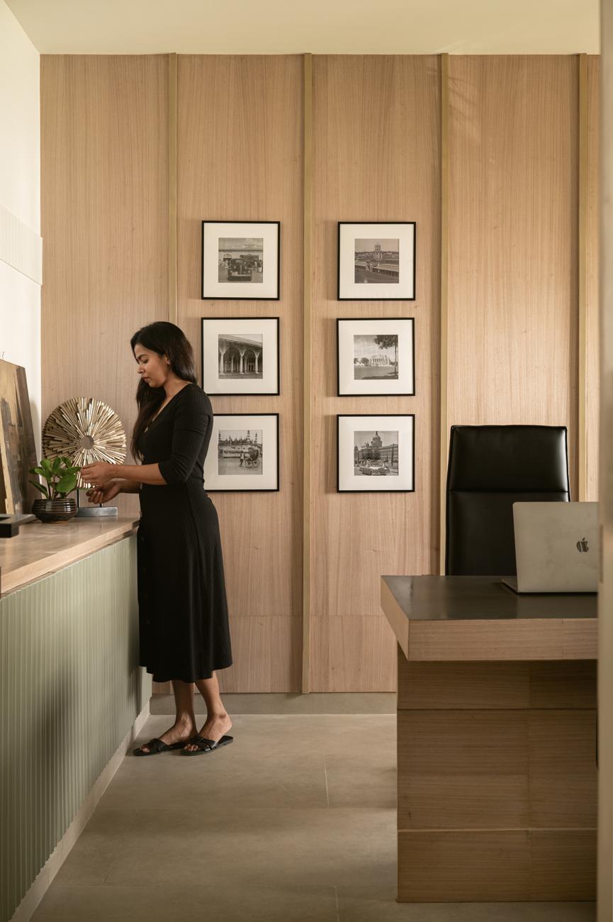 A woman adjusting a brass sunburst sculpture on a green fluted cabinet, beside a wood-panelled wall with six framed black-and-white photographs