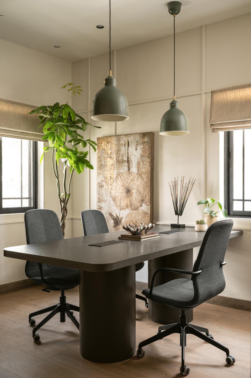 Meeting table with grey chairs under two green pendant lamps, a large textured artwork and a tall plant