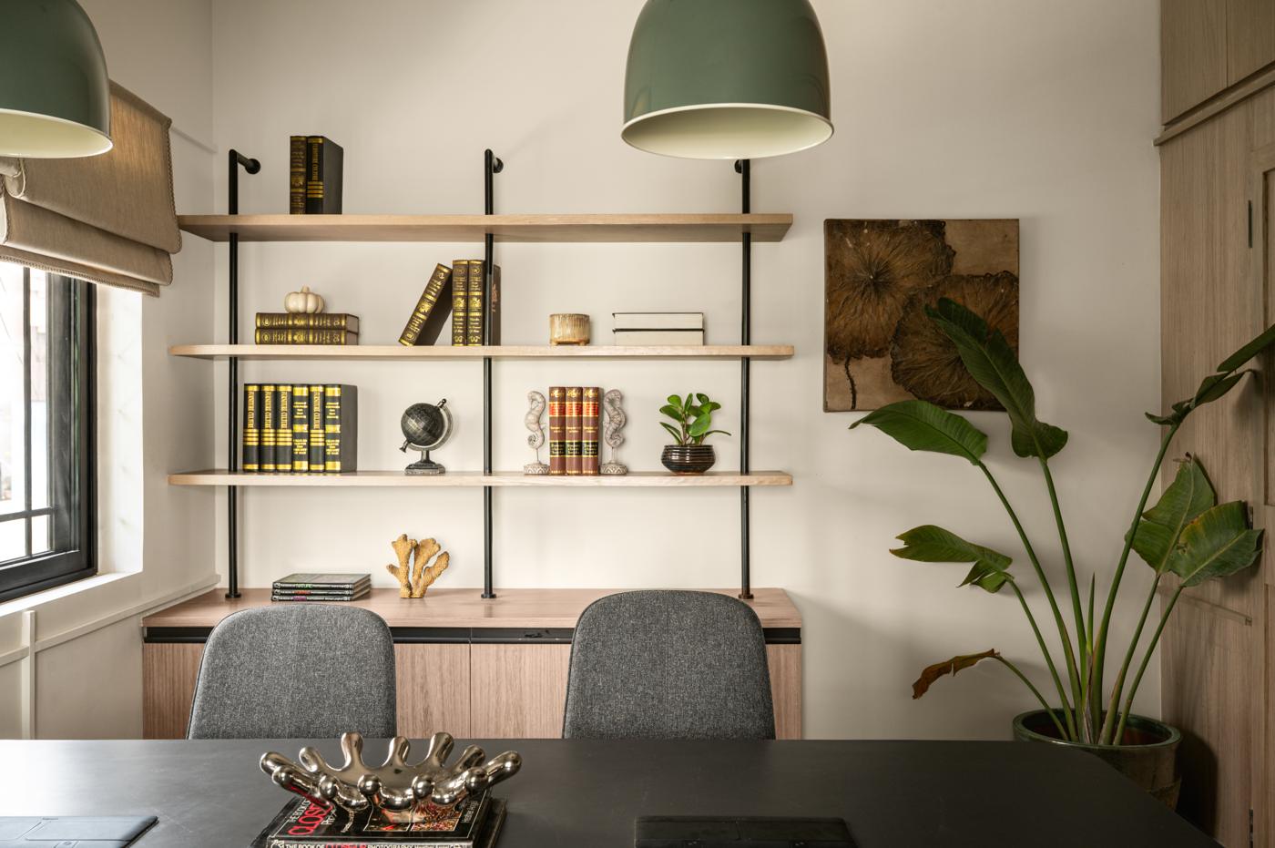 Open shelving with black supports and wood shelves holding books and objects, above a cabinet, with two grey chairs, green pendant lamps and a large plant