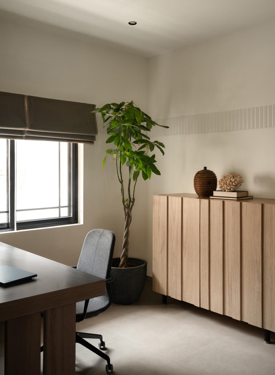 Corner of the office with a plant by a window with a roman blind, a fluted wood sideboard with objects, and the edge of a desk with a grey chair