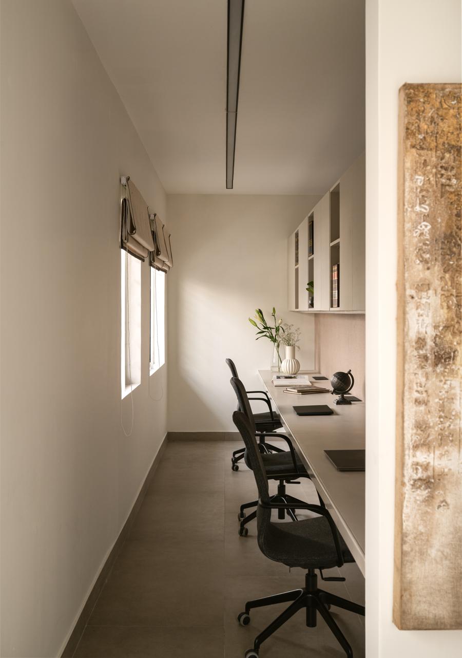 Narrow workspace with a long desk and grey chairs under a linear light, windows on one side and a textured artwork in the foreground