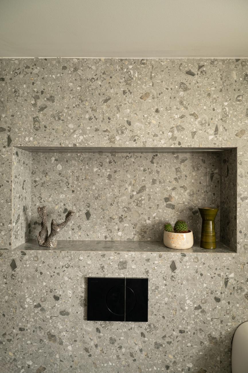 Close view of a terrazzo wall niche holding a coral sculpture, a small cactus and a green vase, above a black flush plate