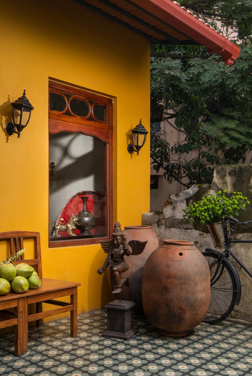 Entry with a yellow wall, a carved timber window, black lanterns, terracotta pots, a bicycle with a flower basket and a patterned tile floor