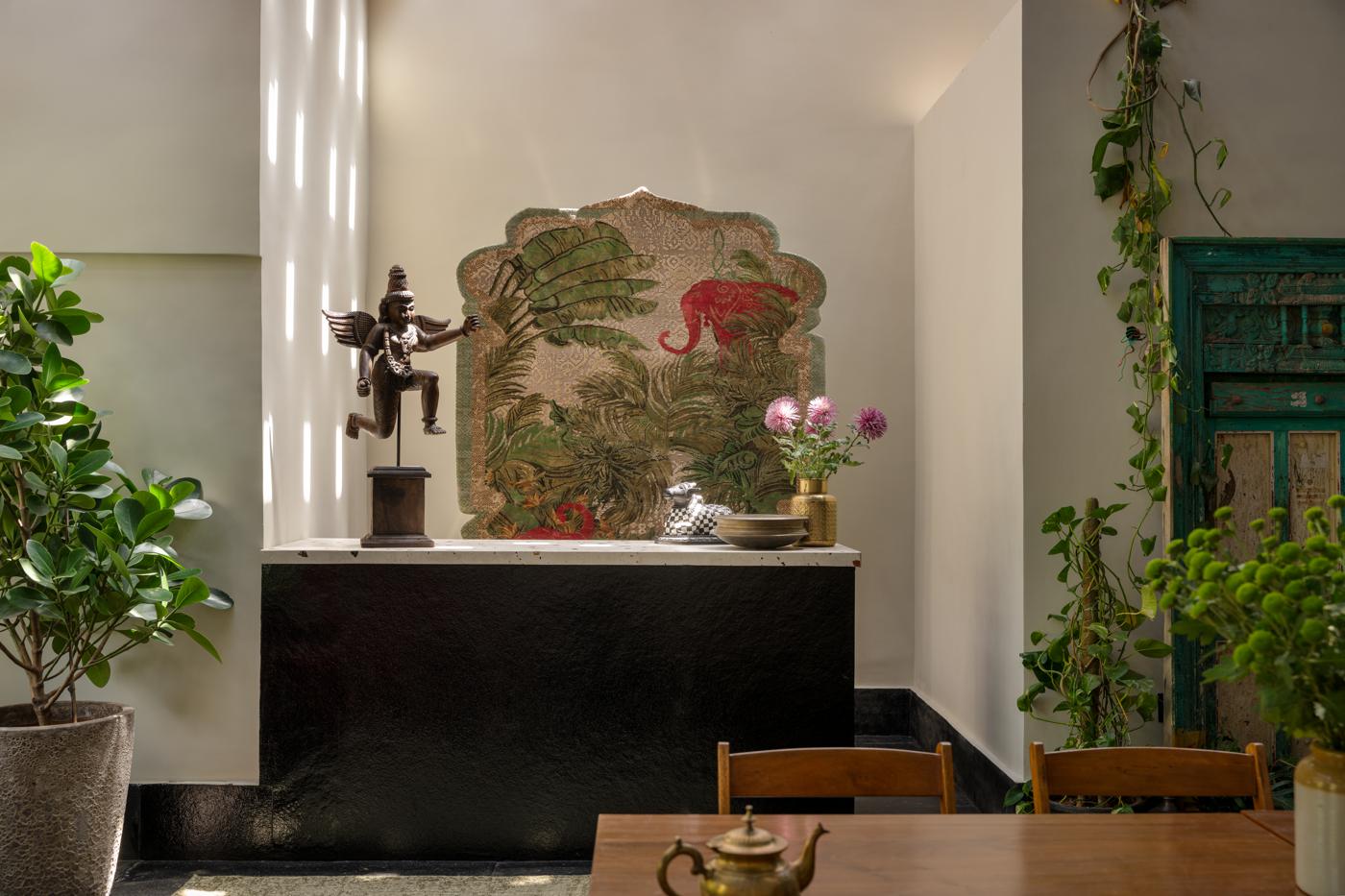 Styled bar counter with a carved figure, flowers and a large artwork, beside plants and a green door