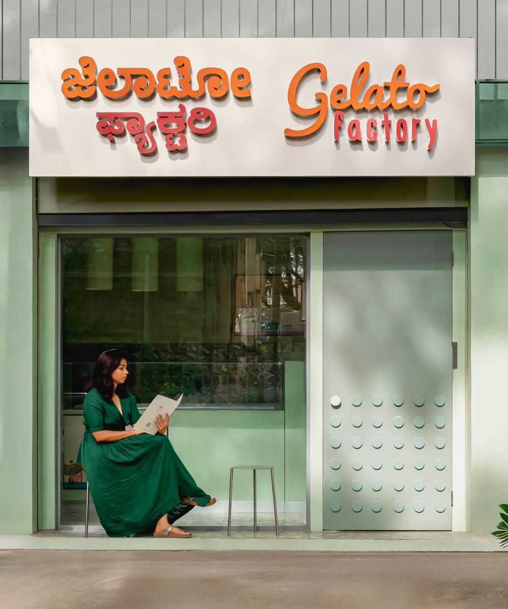 Woman in a green dress reading a menu on a stool inside the glazed shopfront, beside the disc-studded steel door