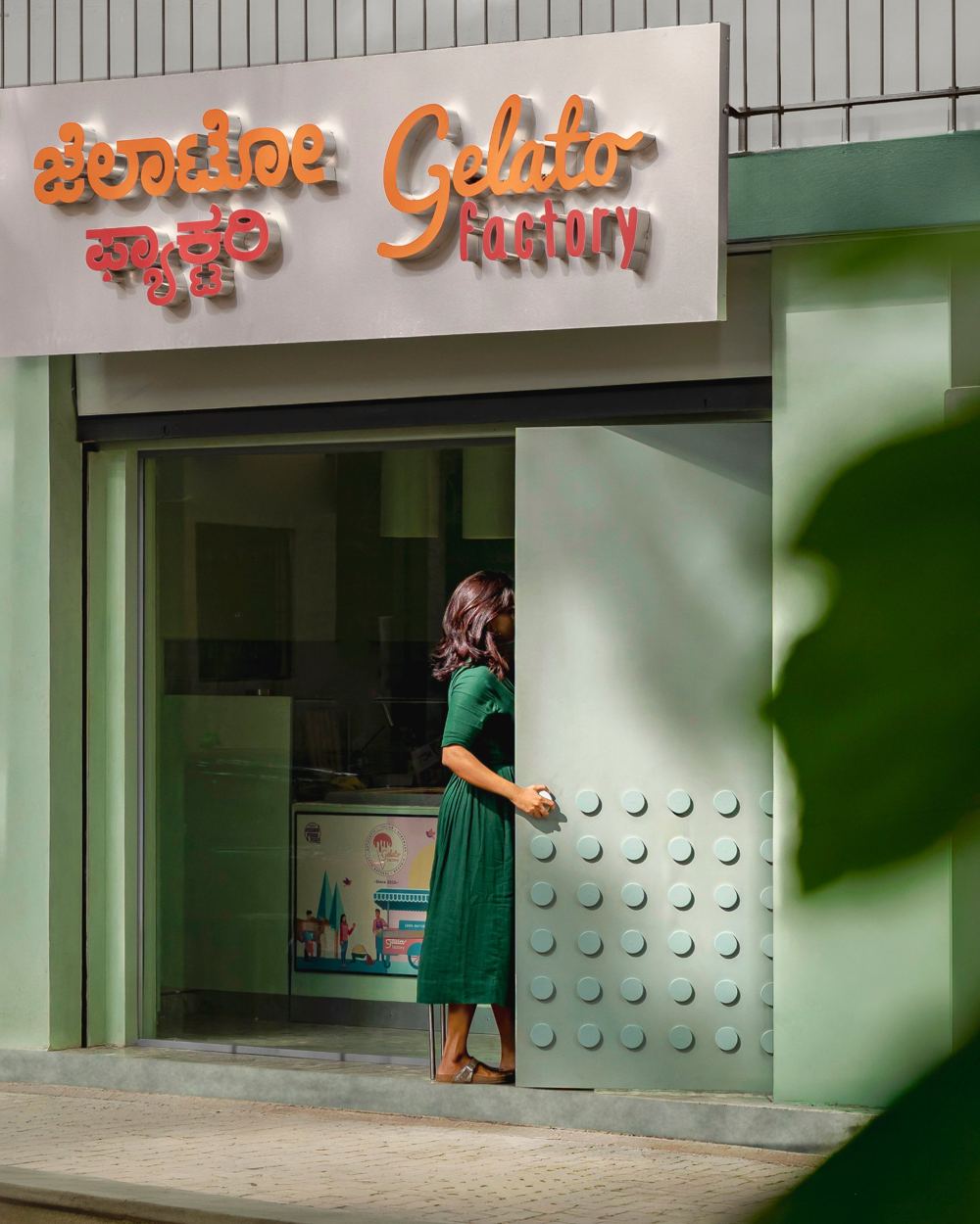 Gelato Factory storefront with a mint-green facade, a steel door studded with raised discs, and a woman in a green dress stepping through, seen through a leaf