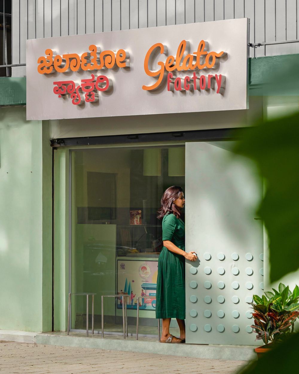 Mint-green storefront with the Kannada and English Gelato Factory sign above a woman in a green dress holding the disc-studded steel door