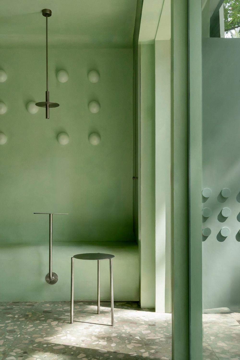 Wall-mounted steel table and a steel stool in front of a wall of domed lights, with a pendant above and the studded door at the right