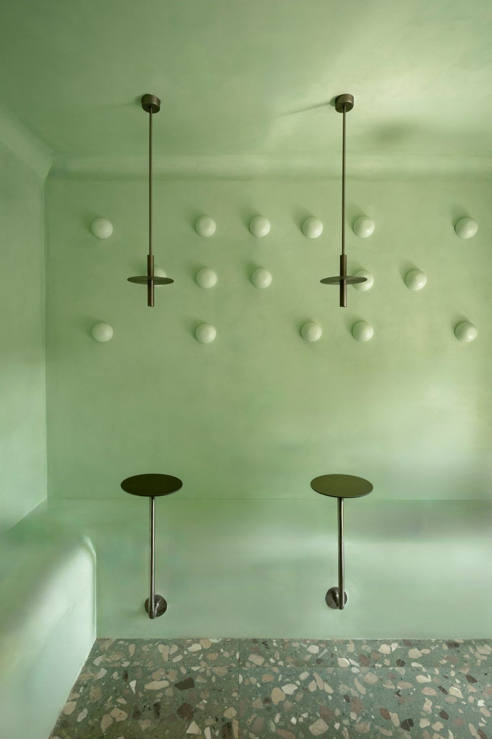 Two bronze pendants and two wall-mounted steel tables against a mint plaster wall of domed lights, above a terrazzo floor