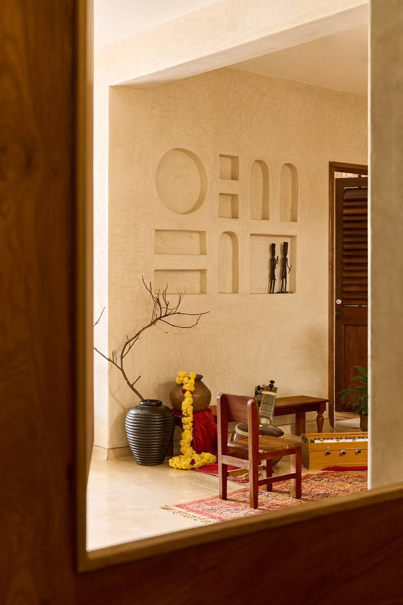 View through a timber-framed opening into a plastered wall of sculpted niches, with a sitar, a harmonium, marigolds and a bare branch in a black vase