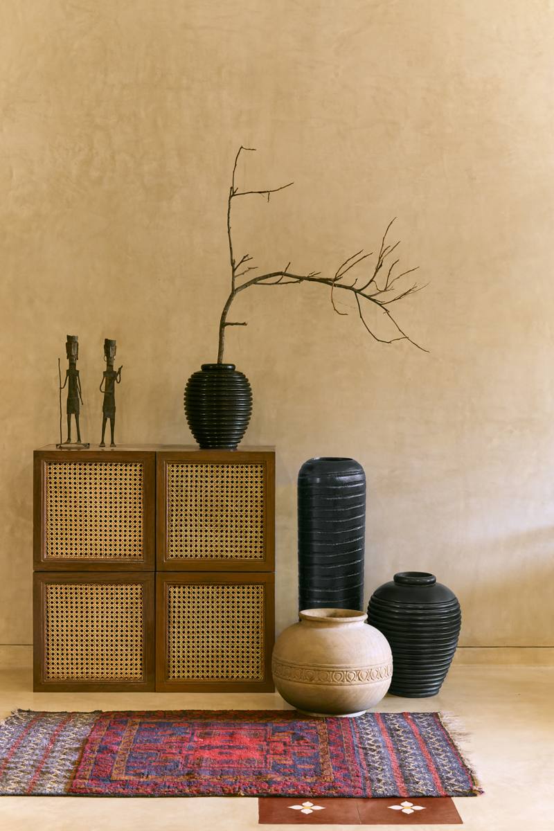 Cane-fronted cabinet topped with bronze figures and a black ribbed vase with a branch, beside carved pots and a woven rug on a plaster floor