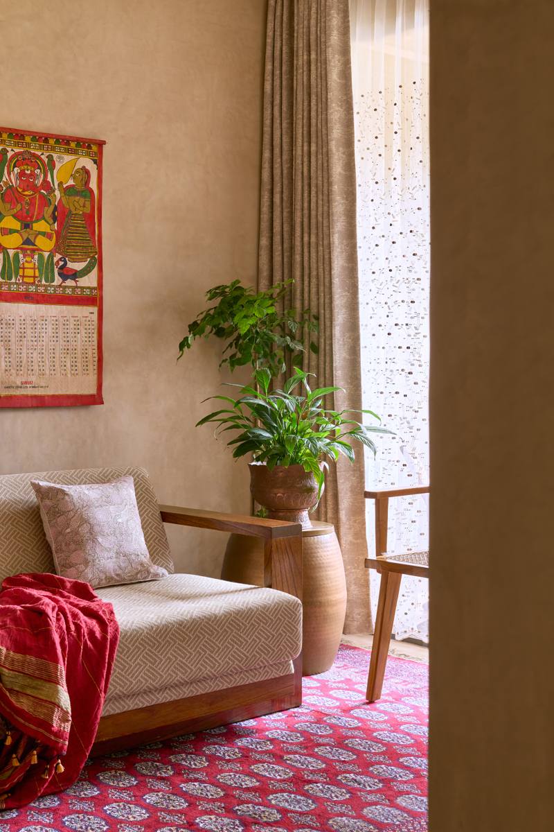 Living room corner with a woven sofa with timber arms, a vintage calendar print, plants in brass pots, sheer and linen curtains and a red patterned rug