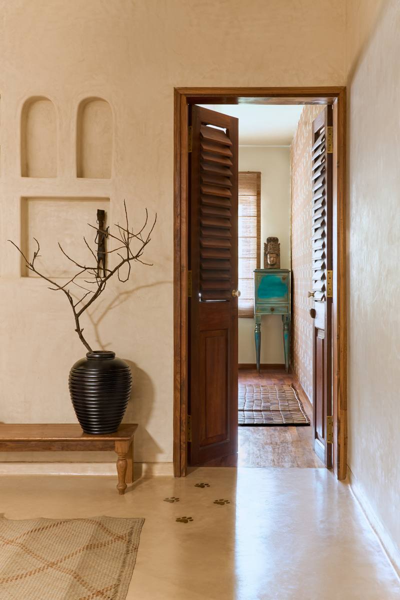 Open louvered timber door leading to a bedroom passage with a teal cabinet and a carved head, beside a black ribbed vase and a branch