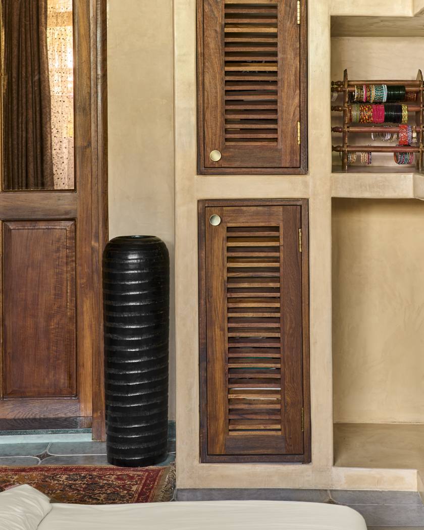 Wider view of louvered timber cabinet doors set in plaster with shelves of bangles, a tall black vase and an antique mirror door