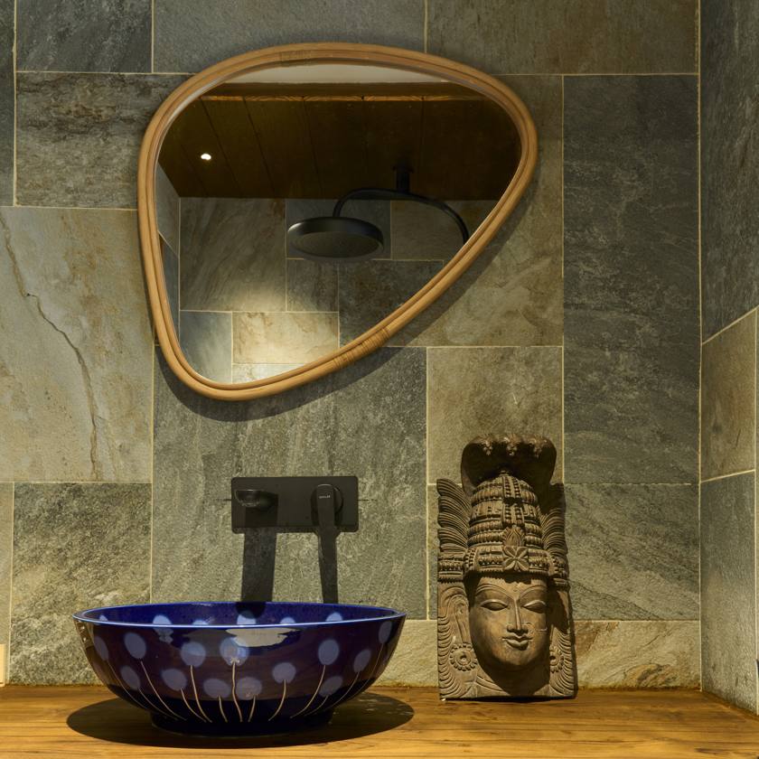 Bathroom with grey stone walls, a teardrop-shaped cane-framed mirror, a blue hand-painted basin and a carved stone head