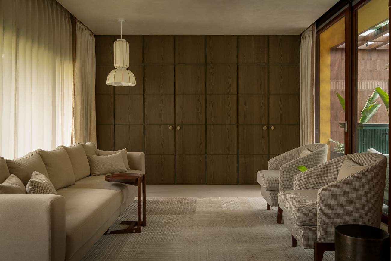 Sitting room with a long linen sofa, tweed armchairs and a pleated pendant light before a closed wall of oak panelled doors with round brass knobs, sheer curtains at the left and a sliding door to the terrace at the right