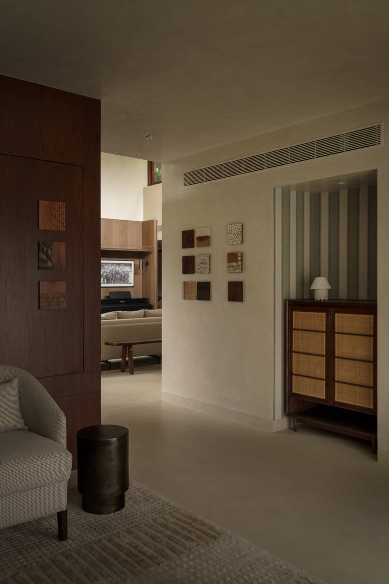 Passage with a grid of carved wooden wall tiles, a striped alcove with a cane-fronted cabinet and a view through to the living area and piano