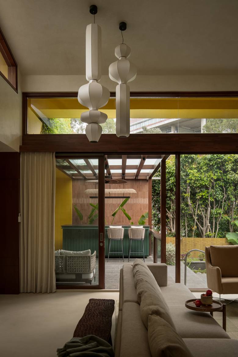 View through the living room to a covered terrace with a green fluted bar, yellow walls and garden beyond, under tall fabric pendants