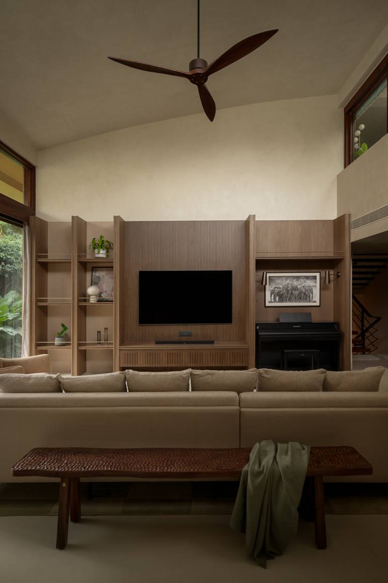 Living room seen from behind the sofa, with a ribbed timber media wall, open shelving with a small white vase, a piano nook and a ceiling fan under a high ceiling
