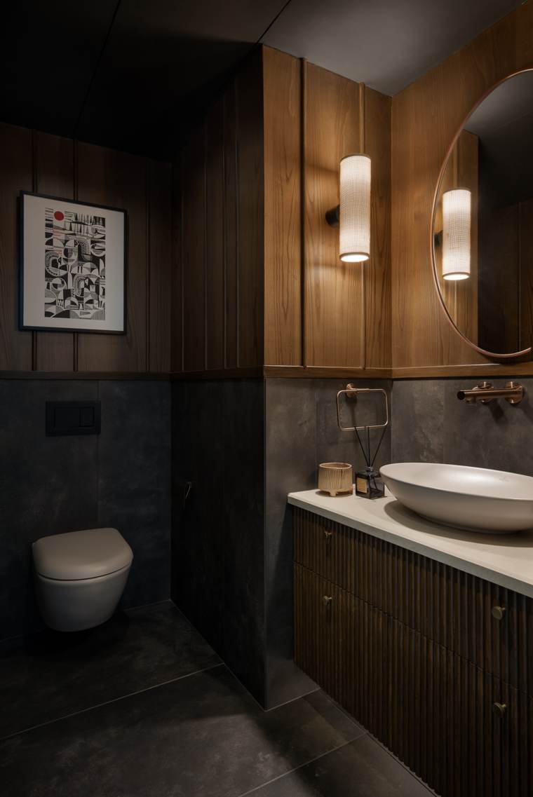 Powder room with timber panelling above dark grey tiles, a cane cylinder wall light, a round copper-framed mirror, a fluted vanity with a white vessel basin and a graphic print