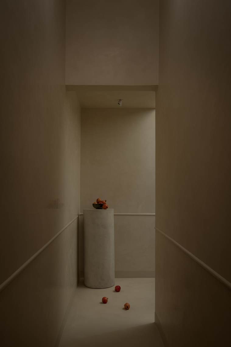 Dim plastered passage with a pale cylindrical plinth holding a bowl of pomegranates, with three more pomegranates on the floor