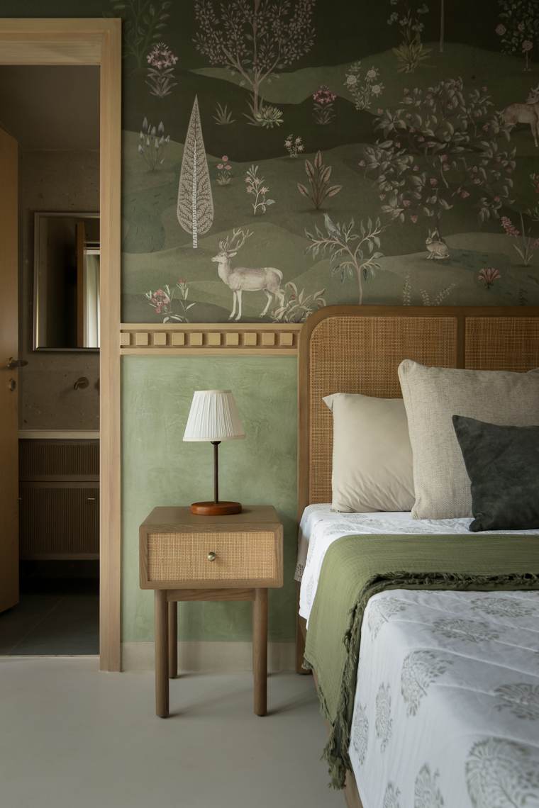 Cane-fronted bedside table with a pleated lamp beside a cane headboard, below woodland wallpaper with a stag, and a bathroom doorway on the left