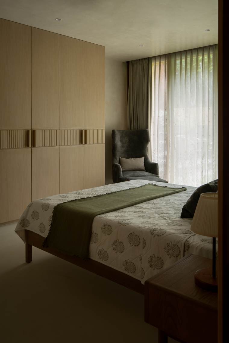 Bedroom with a tall oak wardrobe with fluted handle detail, a grey wingback chair by a sheer-curtained window and a bed with a block-print quilt and green throw
