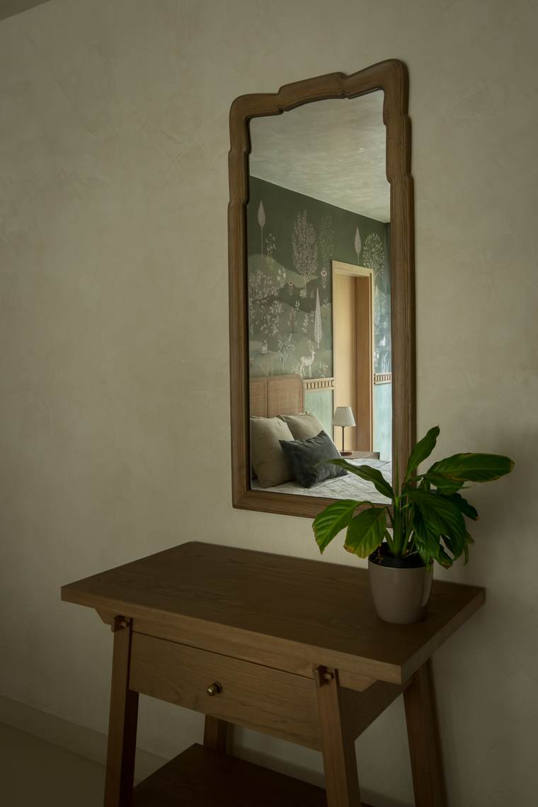 Carved timber-framed mirror above a slim timber console with a peace lily, reflecting a bedroom with woodland wallpaper
