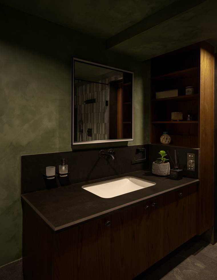 Bathroom vanity with a black stone counter and white basin, wall-mounted tap, framed mirror and timber shelves on olive plastered walls